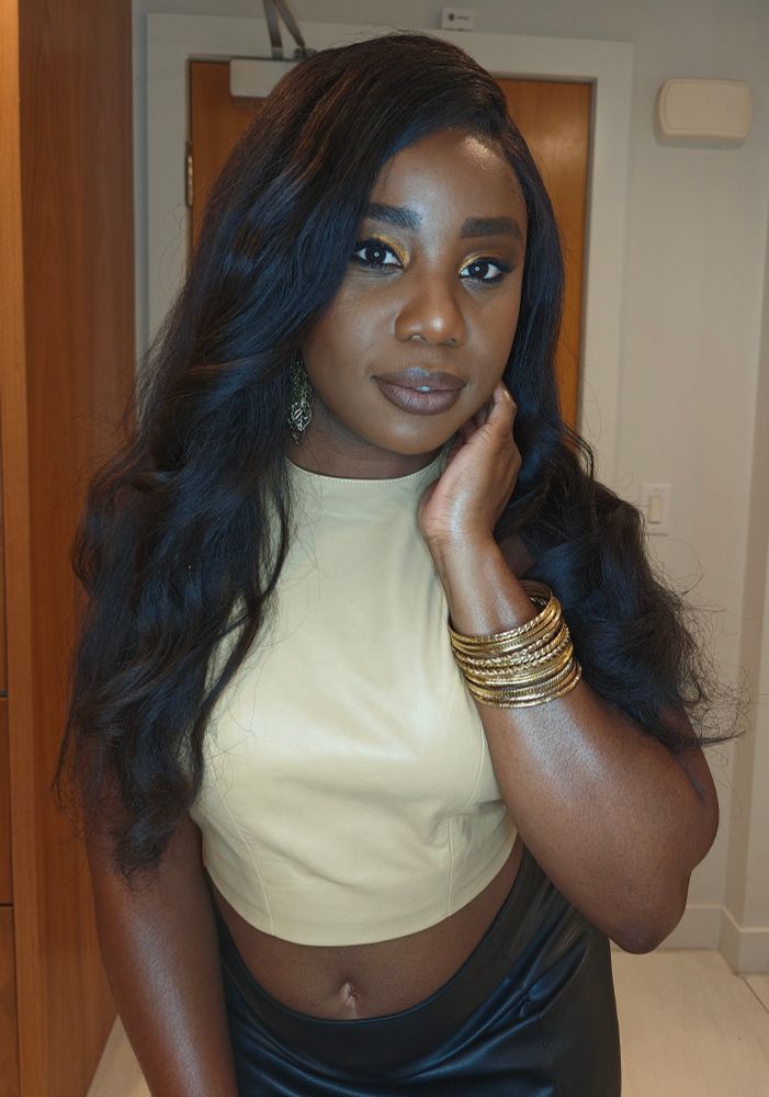 A Black woman wearing a tan leather top and black leather skirt looks directly into the camera. She has her hair styled in loose curls that have been brushed out. She is also wearing gold bangles on her right hand, which is placed on her neck. 