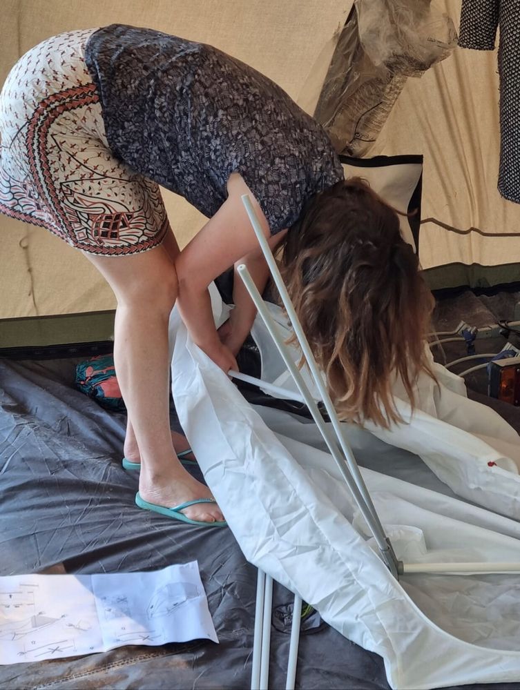 Woman bending over, busy putting up a complicated pop up ikea wardrobe in a tent at a camping site.