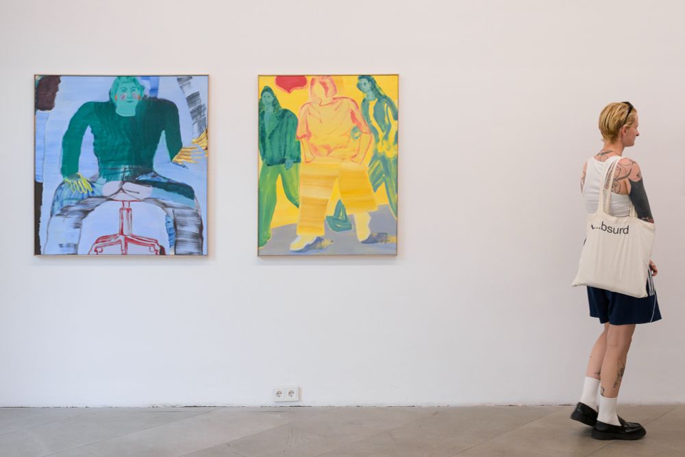 Eine blonde Person mit kurzen Haaren, einem weißen Tanktop und einem Stoffbeutel mit dem Aufdruck ‚…bsurd‘ betrachtet ein Gemälde. Zwei Kunstwerke von Lisa Breyer sind zu sehen, das rechte zeigt drei stilisierte, grüne und gelbe Figuren vor einem gelben Hintergrund, das linke zeigt eine abstrahierte Figur, sitzend.