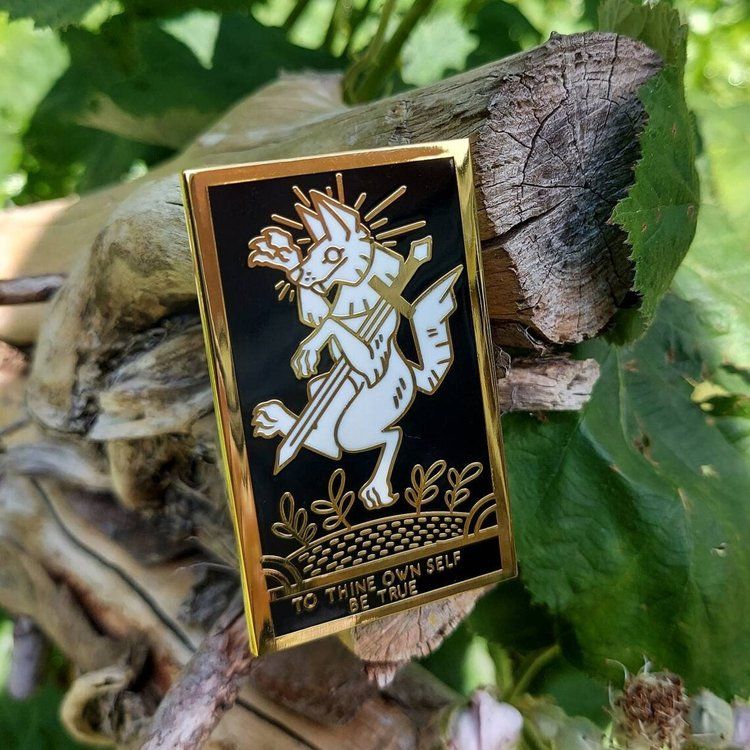 A photo of an enamel pin featuring a heraldry styled white wolf that's clutching a sword. Under the image are the words To Thine Own Self Be True