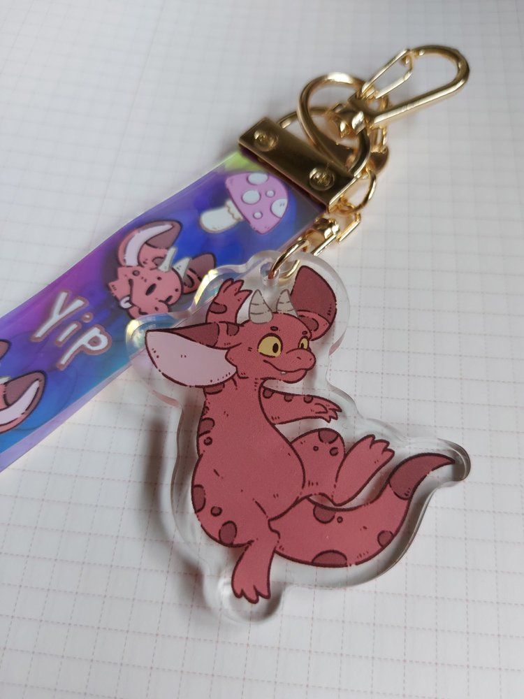 A photo of a red kobold acrylic charm attached to a holographic PVC lanyard that has little kobold faces a mushroom and the word yip on it.