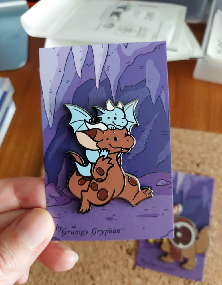 Photo of an enamel pin featuring a kobold carrying a blue dragon whelpling on their shoulders.