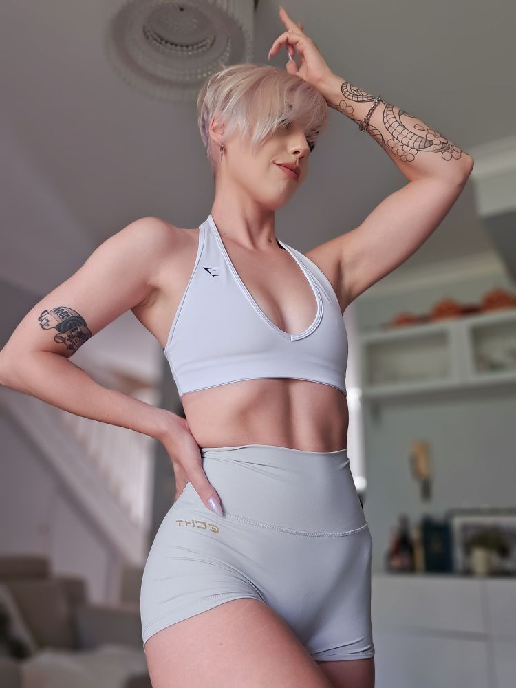 A photo of Bonez posing with one arm up above their head and the other hand resting on their cocked hip. They're wearing a white halterneck top and a pair of stone grey shorts. 