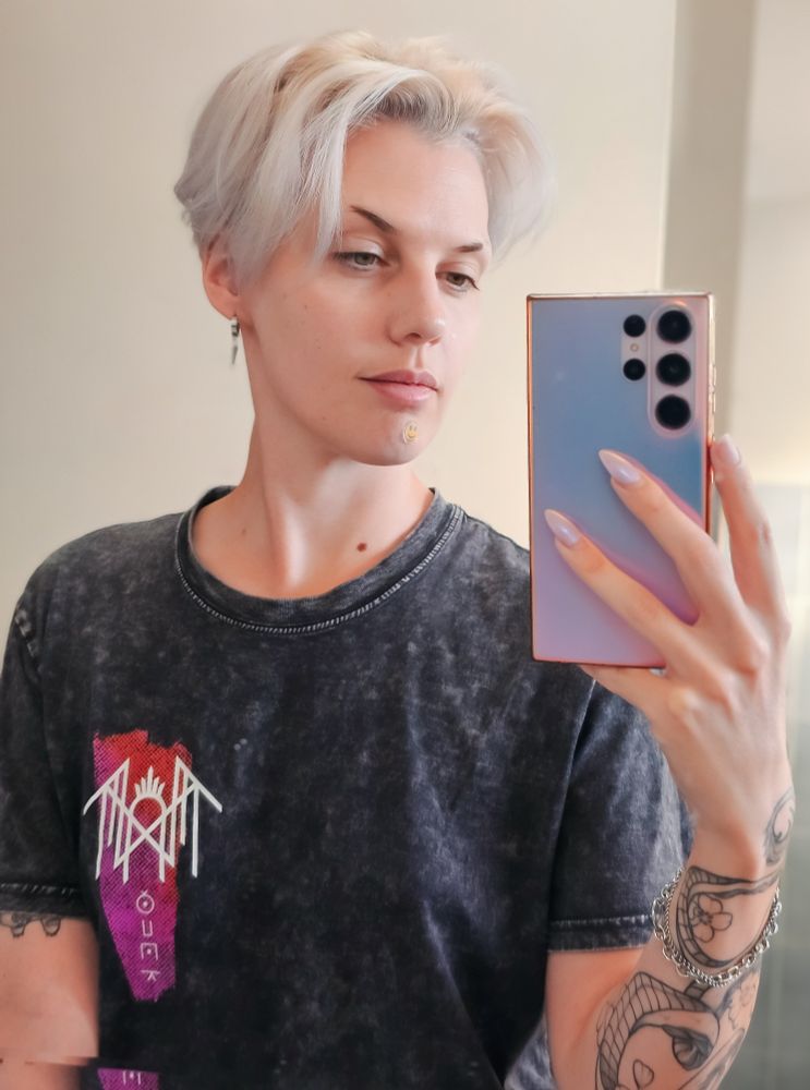 A mirror selfie of Bonez. They are fresh faced, wearing a distressed-looking dark grey sleep token tshirt and their hair is parted in the middle. 