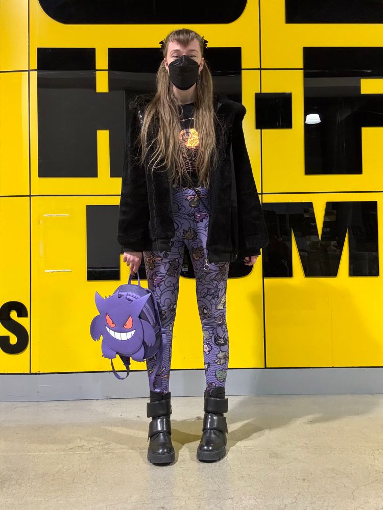 iz, a person with long blonde hair and a black mask, stands like a statue against a yellow wall, there’s high contrast with the wall against their purple leggings covered in ghost pokemon, their bright purple Gengar shaped backpack dangling from their right hand, and their big black fluffy coat and tall boots. 