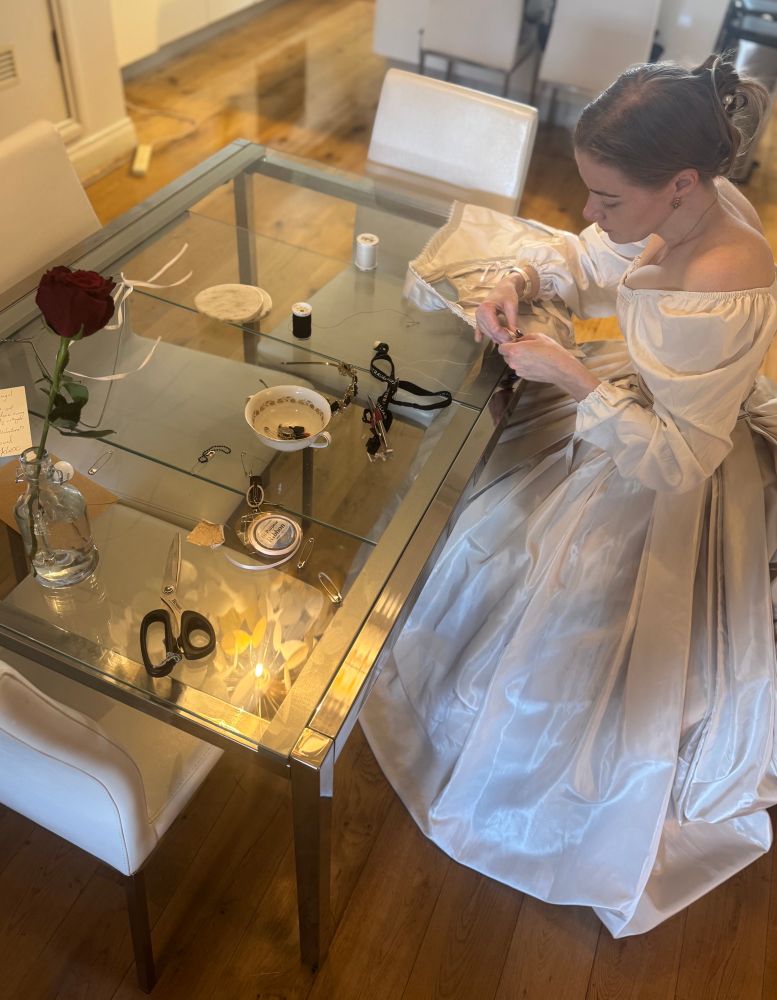A warm toned photo from the left side of Iz, a femme individual, sitting at a glass table hand-sewing a corset cover. She is perched on the edge of a chair in a large taffeta skirt and cage crinoline underneath, and is wearing a corset and chemise. Tools litter the table - ribbon, scissors, safety pins, a pair of snips on a lanyard, a teacup with a thimble in it - as well as a single red rose for Valentine’s Day.

