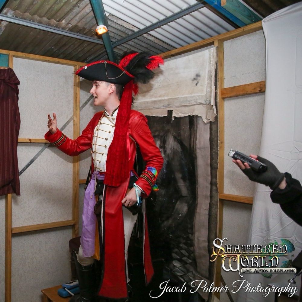 A LARPer in a fabulous red greatcoat made of genuine leather stands orating in a tavern. The coat has a false front made of white leather with gold soutache braid, with a matching stand collar edged in gold and black braid. Black and gold trim sewn in loops on the long sleeves indicate this character is an Admiral. 

The photo is credited to Shattered World LARP and Jacob Palmer Photography. 

Iz made the greatcoat to her bespoke measurements! 