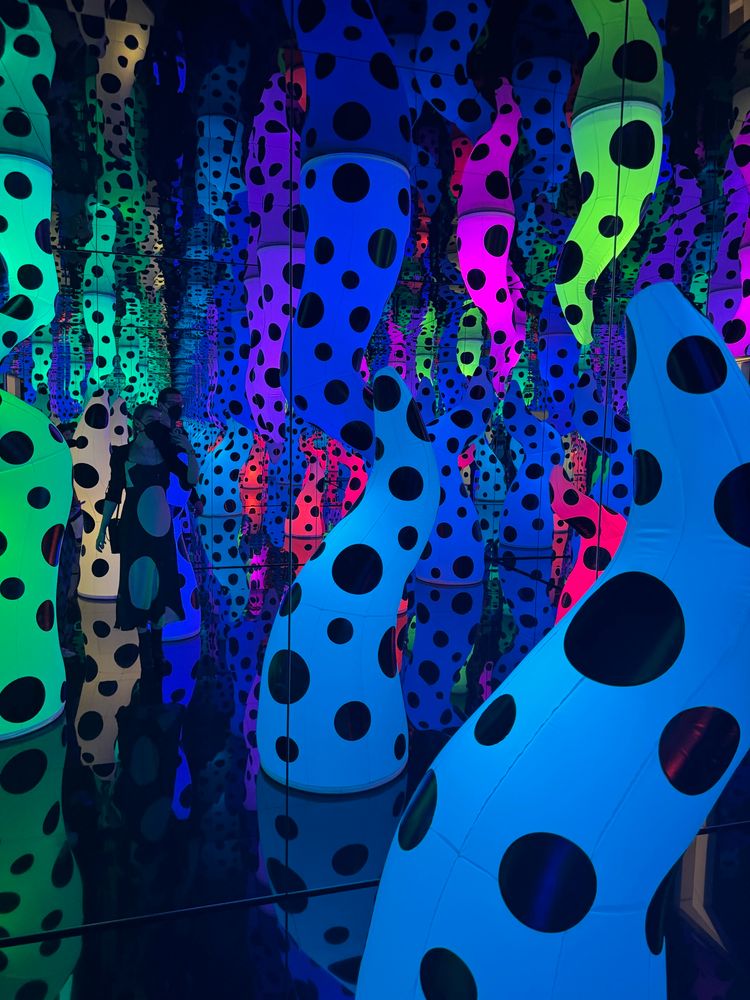 A technicolor fever dream - this mirrored Yayoi Kusama infinity room is full of inflatable tentacle shapes, lit from within in various colours, predominantly blue, and covered in black polka dots. The effect of the mirrors makes it appear the tentacles go on forever. A femme person in a black mask and a black dress with large white spots takes a photo in the mirrored wall to the left of frame, though she appears small compared to the scale of the tentacles. 