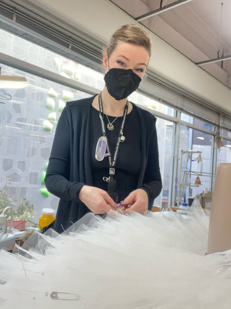 Iz, a person wearing a black mask and dressed in all black to match, stands smiling at the camera from behind a leg mannequin with an upside down tutu on it. They hold snips in their left hand and thread and an awl in their right, working on the hand shanking process holding all layers of the tutu together. 

The lanyard around their neck reads “The Australian Ballet” and they have a pronoun pin reading “she/they” on it. 