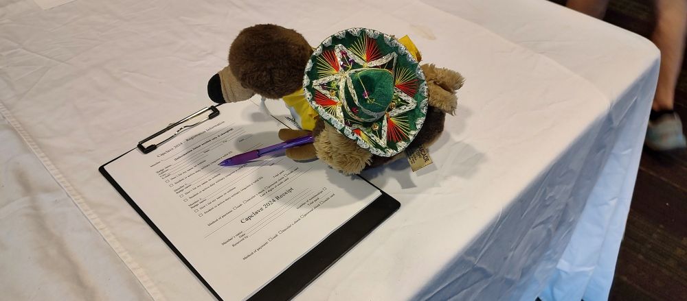 Pedro filling out the Capclave 2024 registration form, he's so excited about next year. He is wearing his usual yellow and white shirt with a green sombrero. He holds a pen with purple ink in his left claw because purple ink makes everything better. Did we mention that Capclave memberships make great winter solstice related holiday gifts?
