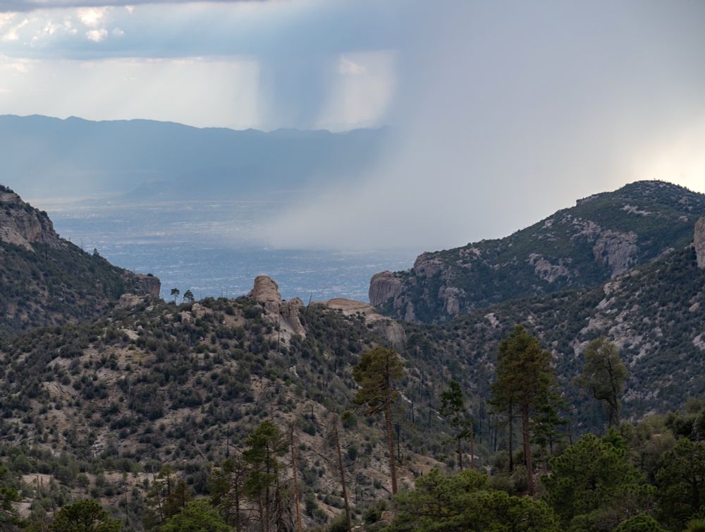 A large column of rain with sun highlighting the right side and the left in shadow hits a valley below a ridge of rocks and trees in Tucson Az 