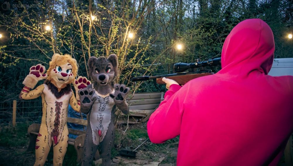 Xei and Akamaru are in bodypaint and fursuit with their paws up in the air. A rifle is being aimed at them by a hunter in a pink hoodie. They’re outside by the woods