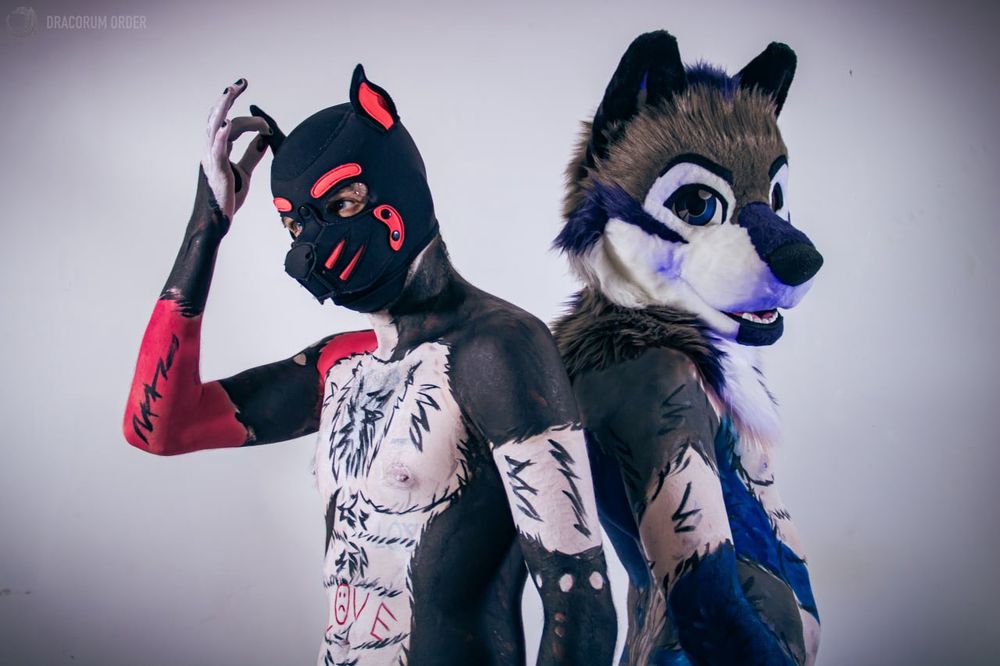 Scruffy and Smudge in bodypaint back to back. The former is wearing a black and hot pink pup hood fiddling their right ear. The latter is in a fursuit head looking at the camera