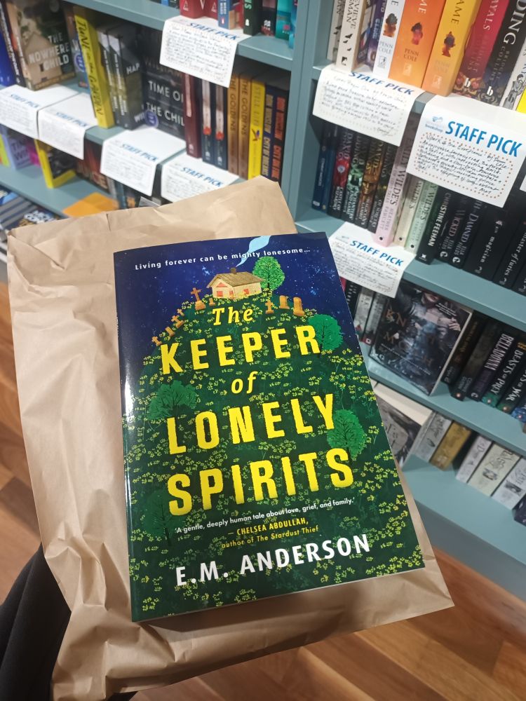 A photo of someone holding the book The Keeper of Lonely Sprits by E.M. Anderson. In the background are the sci fi/fantasy shelves of a bookshop.
