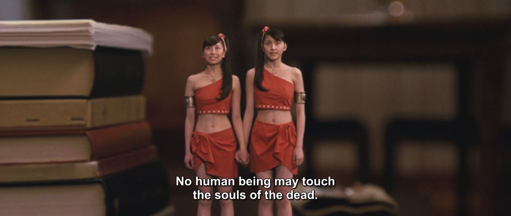 A frame from Godzilla: Tokyo S.O.S. (2003).
Two similar (but not identical) women in matching red two piece outfits stand next to each other clasping hands. There is a nearby stack of books that is the same height as them.
The caption reads, "No human being may touch the souls of the dead."

Not pictured: Godzilla; g-g-g-ghosts