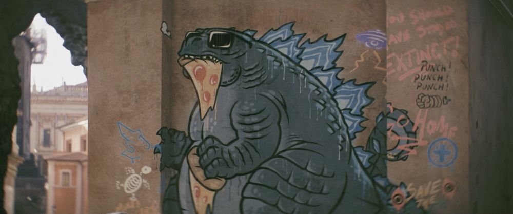 A frame from Godzilla X Kong: The New Empire (2024).
Graffiti of Godzilla on a brown stone wall outside. Godzilla in this wall art is more squat, round - one could say he is a chonker - and he looks so damn cool here. He's got sunglasses on and he's eating two slices of pepperoni pizza at once. He looks like a fun hang but also would have no problem calling anybody out for being an asshole. He's like fun uncle Godzilla that likes bowling and buys beer for you

Other bits of graffiti around Godzilla include a flying saucer that looks like it's from "Invasion of Astro-Monster", a skeletal turtle (likely a reference to the turtle seen in "Mothra vs Godzilla"), and Hedorah's eyes.

There are also some words such as "Go Home" and "You Should Have Stayed Extinct!" which probably should have been written in Italian since this is in Rome but whatever.

Not pictured: Kong; a new empire (Rome is literally an old empire)