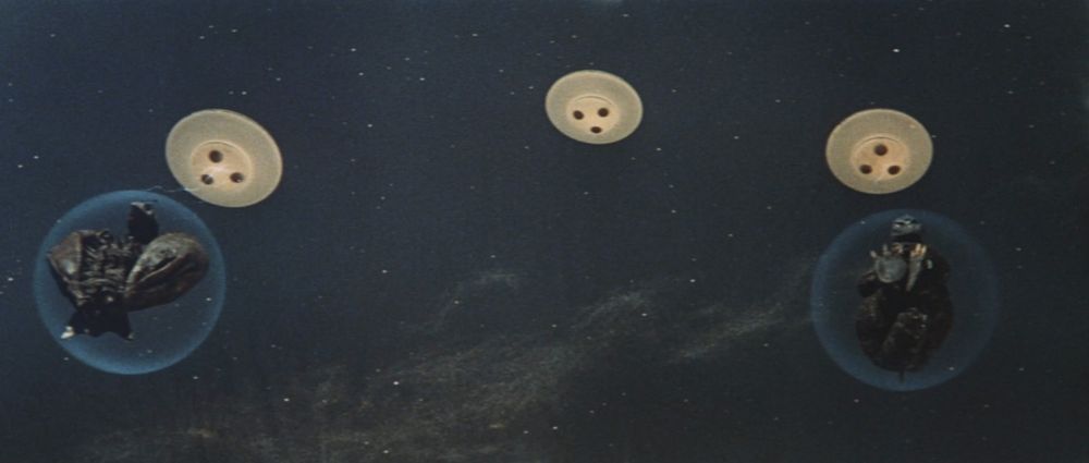 A frame from Invasion of Astro-Monster (1965).
We see a vast dark sky filled with stars. On this cosmic canvas, three flying saucers. Two of the interstellar craft are carrying blue bubbles in tow. Inside these bubbles are Godzilla and Rodan, all curled up.