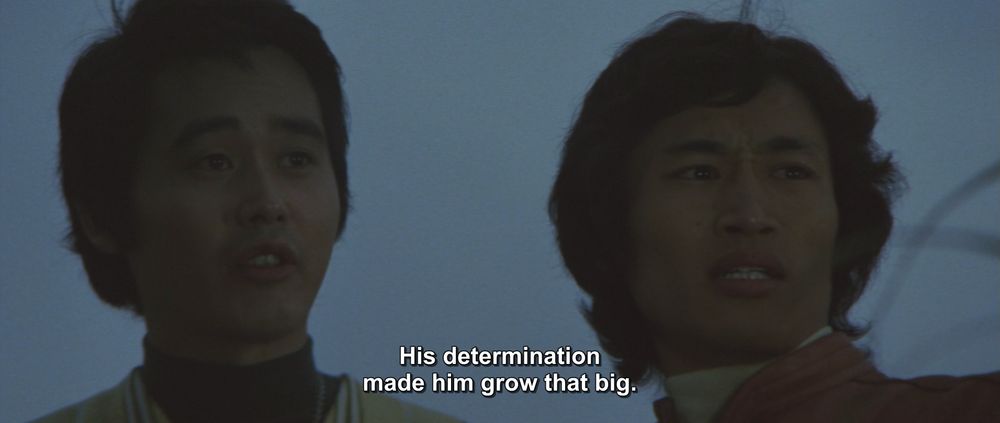 A frame from Godzilla vs Megalon (1973).
Two men stand outside, it looks like day is maybe just about to break. They are looking off to the side with expressions of amazement. 
The caption reads, "His determination made him grow that big."

Not pictured: Godzilla; Megalon; whoever is offscreen cultivating mass