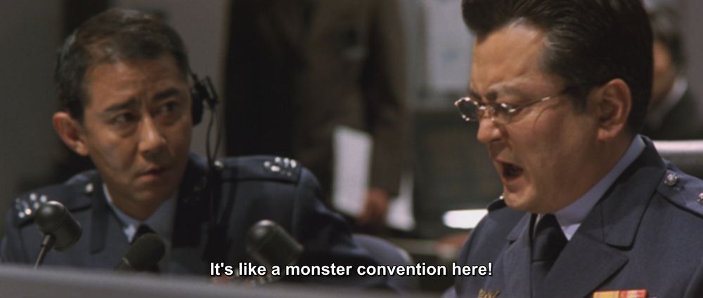 A frame from Godzilla, Mothra, and King Ghidorah: Giant Monsters All-Out Attack (2001), AKA GMK.
Two military-ish men are sitting at a console. One is wearing a headset and looking at the other, who is expressing some outrage in the current moment. 
The caption reads, "It's like a monster convention here!"

Not pictured: Godzilla; Mothra; King Ghidorah; AKA the monster convention