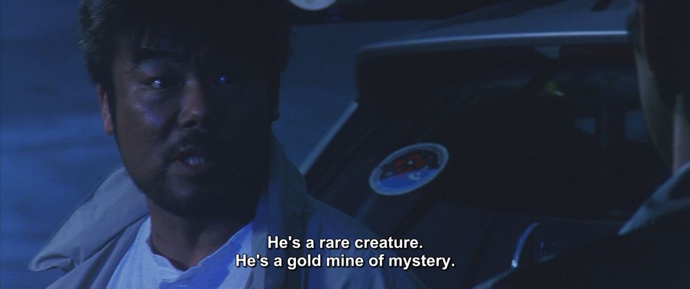 A frame from Godzilla 2000 (1999).
A man wearing an open jacket stands outside, just behind a car. He is looking at another man who is in the foreground, just barely in the frame. It looks like its early morning or late evening since everything is bathed in a blue light.
The caption, "He's a rare creature. He's a gold mine of mystery."

Not pictured: Godzilla; that special someone you describe as a gold mine of mystery