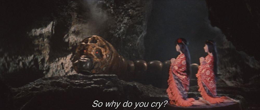 A frame from Ghidorah, the Three-Headed Monster (1964).
Two women in matching red robes kneel in a cave, where a large brown caterpillar with red eyes resides. They seem to be singing to the caterpillar. 
The caption reads, "So why do you cry?"