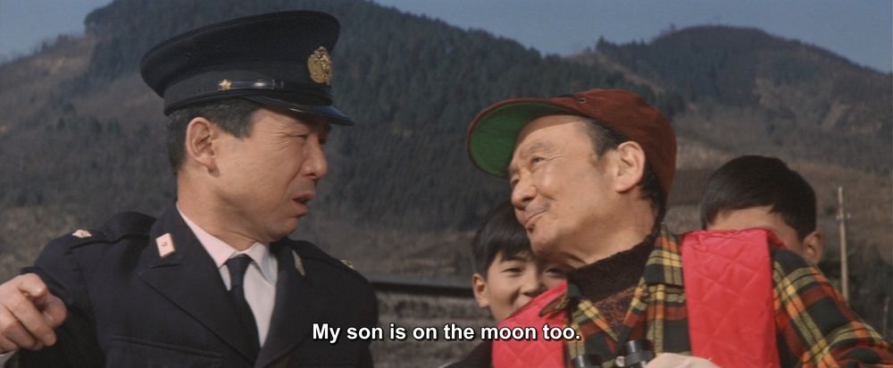 A frame from Destroy All Monsters (1968).
A small gathering of people outside with some small mountains in the background. A police officer is talking to an older man who looks like he's outfitted for hunting or fishing.
The caption reads, "My son is on the moon too."

Not pictured: Godzilla; moon son