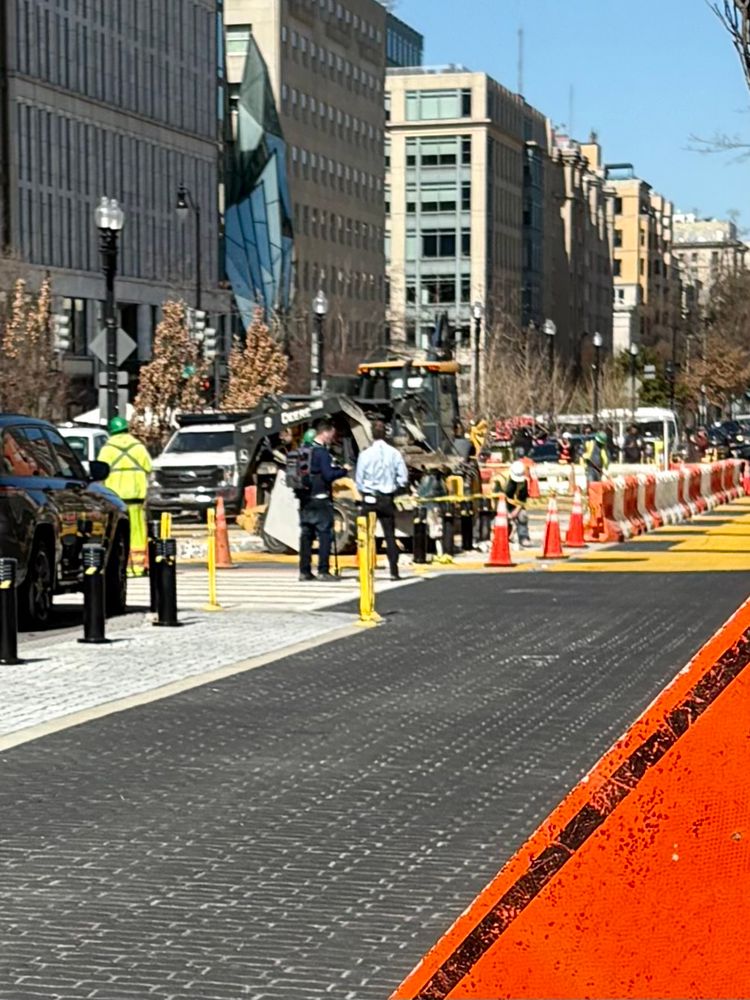 Road work being done as small machinery digs up the road. Around them are warning signs, protesters and news outlets trying to get interviews