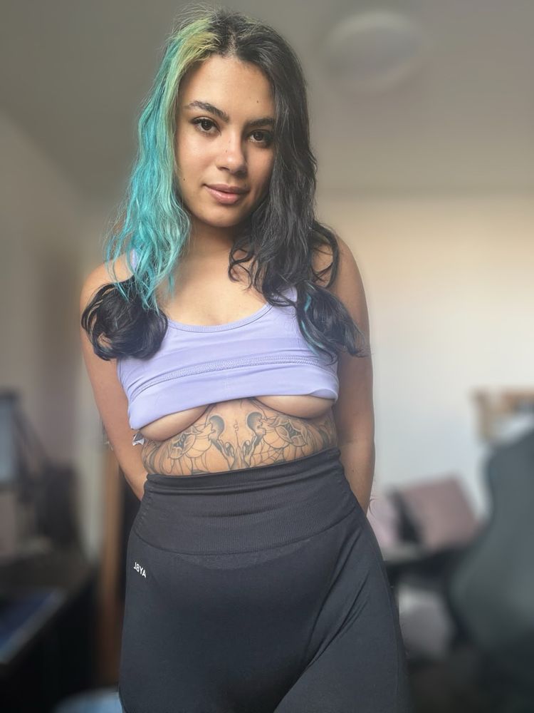 A person is standing in a casual pose, wearing a light purple top and high-waisted black leggings. Their long hair features blue highlights, and several tattoos are visible on their abdomen. The background includes a glimpse of a room with a computer setup, creating a relaxed atmosphere. The lighting is bright, highlighting the details of the outfit and tattoos.