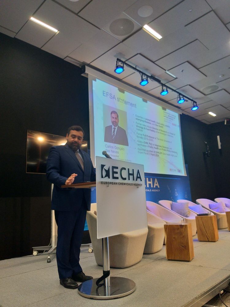 Carlos Das Neves (EFSA's Chief Scientist) standing at a podium giving a presentation at a conference hosted by the European Chemicals Agency (ECHA). Behind him, a large screen displays a slide titled "EFSA statement" with a photo and biography of Carlos Gonçalo das Neves, highlighting his expertise in animal health, food safety, and policy. 