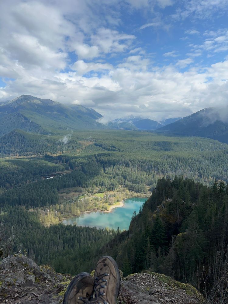 My feet are crossed at the edge of a rocky outcropping overlooking a deep wide green valley at the base of m steep mountains. Far below a light blue lake is hit by sunlight under a bright blue sky dotted with clouds. 