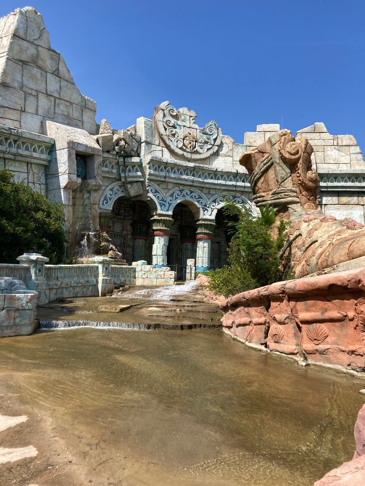 The facade of Poseidon's Fury as seen from the left side of the courtyard along the former attraction's exit path. The facade has a crumbling fracture on its left side, a rift in the ground, and a broken statue in front of it.