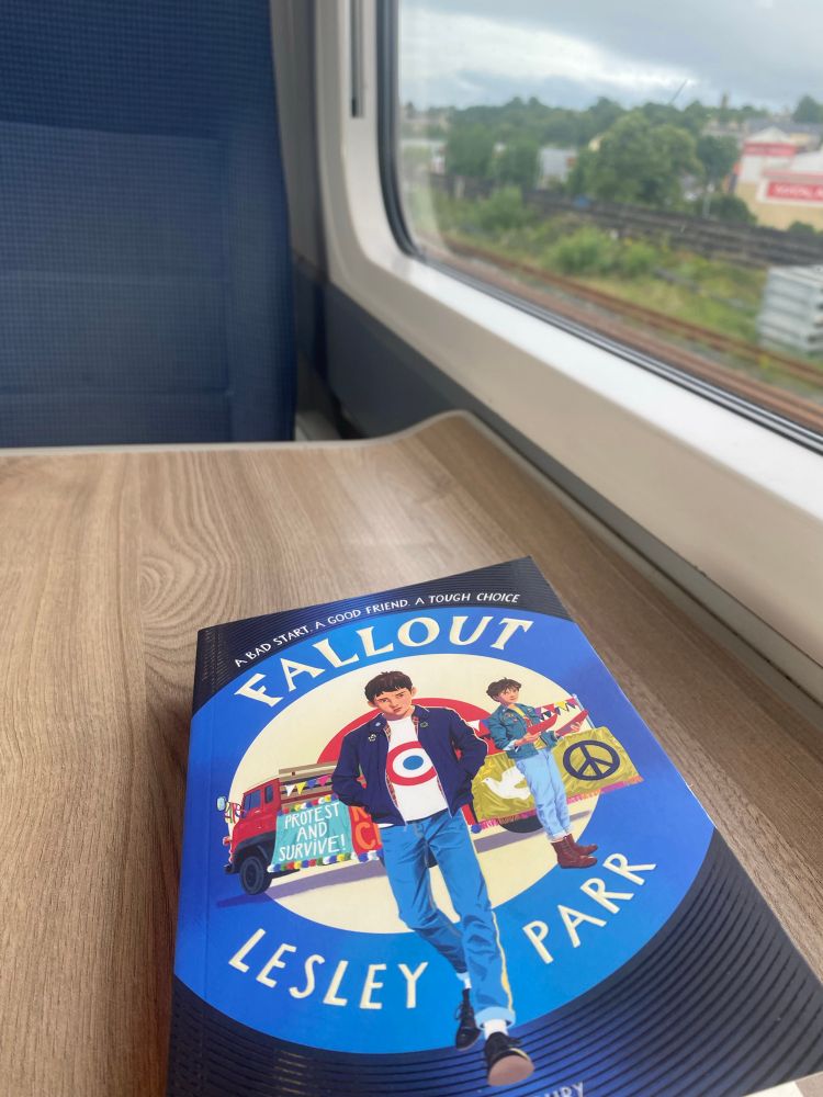 My view from my seat on the train - trees out the window and my book (Fallout by Lesley Parr) in front of me. 