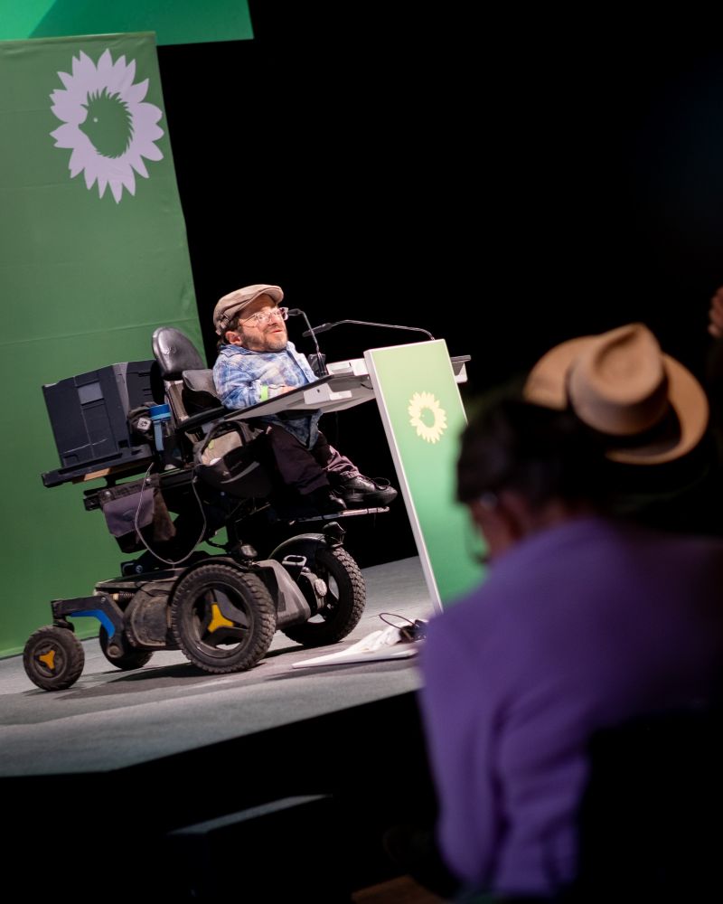 Raul Krauthausen spricht am Redepult des Stadtkongress. Das Redepult und der Hintergrund sind grün gebrandet und eingefärbt. Im Vordergrund Zuhörer*innen.