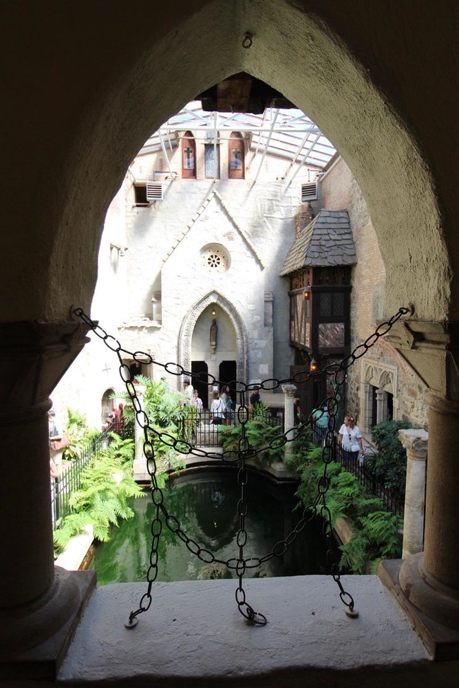 A view through an arched window showcasing an inner courtyard with lush greenery, a reflective pond, and rustic architecture. People can be seen exploring the area, surrounded by stone and timber structures under a partially glassed roof.
