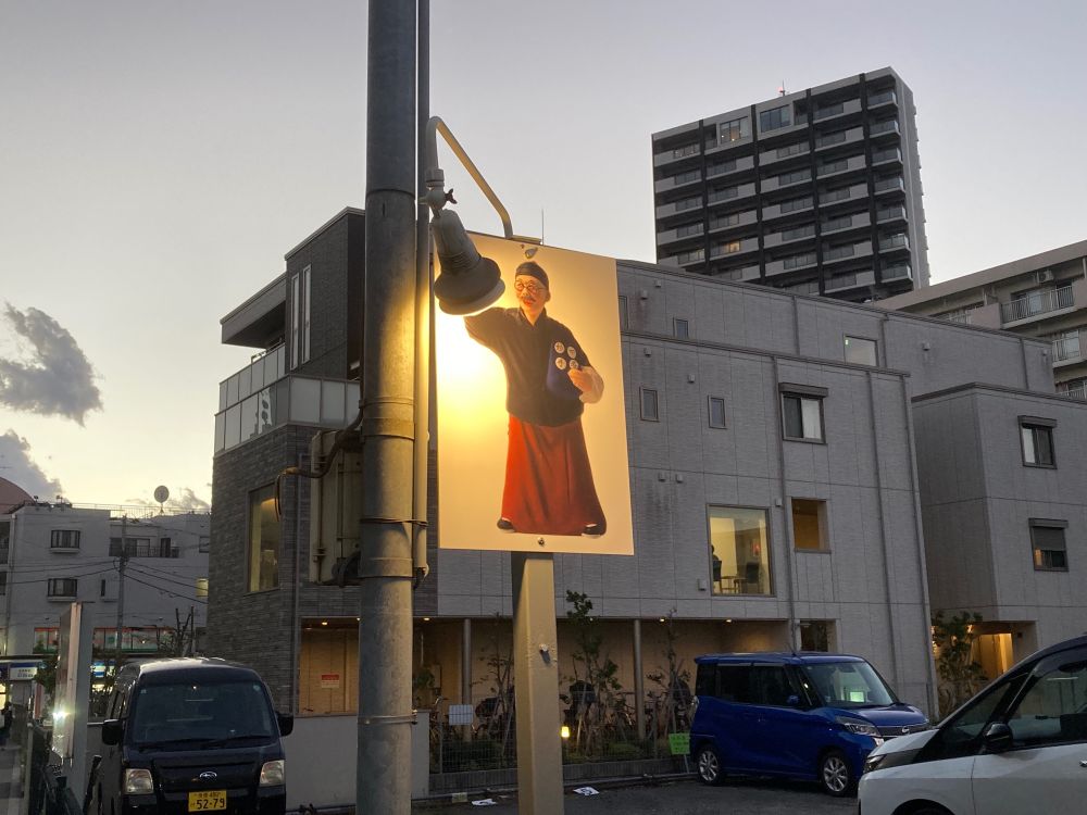 A street scene at dusk featuring a brightly illuminated sign of a character in traditional clothing, holding something in one hand. Surrounding buildings are modern, and parked cars are visible in the foreground.