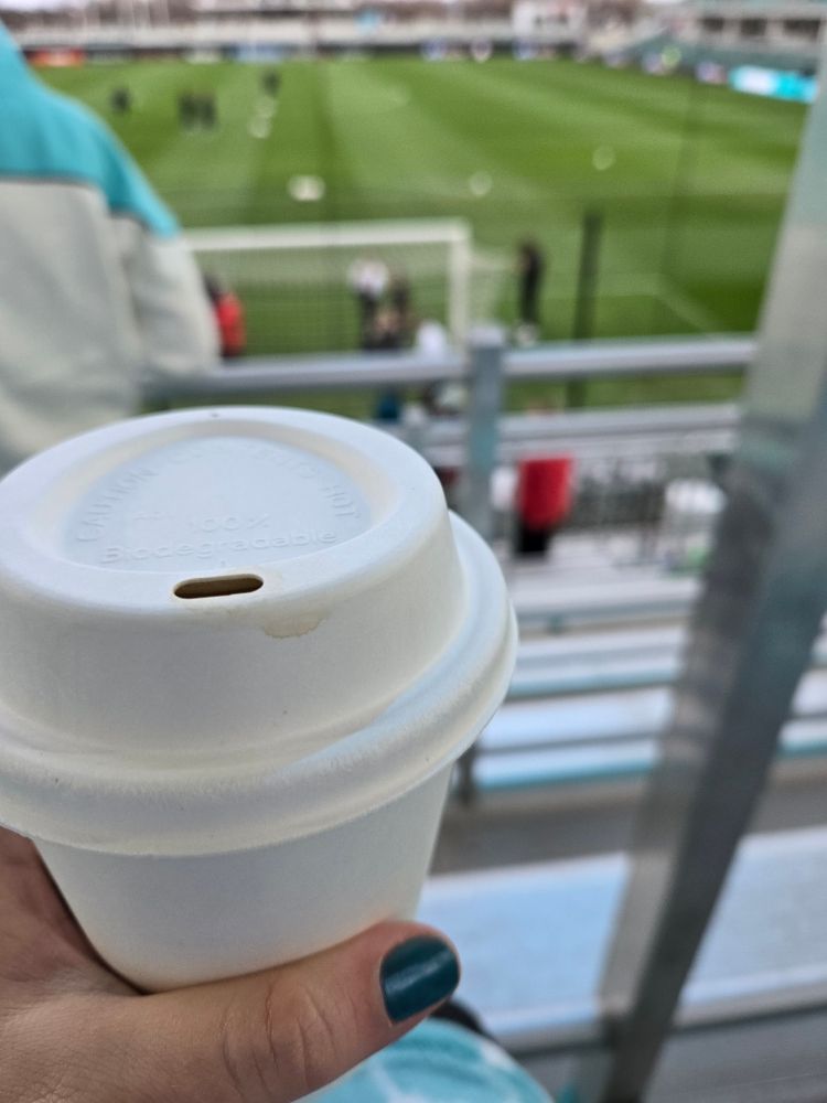 Picture of a hand holding a coffee cup. The thumb nail is painted teal. In the background, out of focus, is the pitch at CPKC stadium