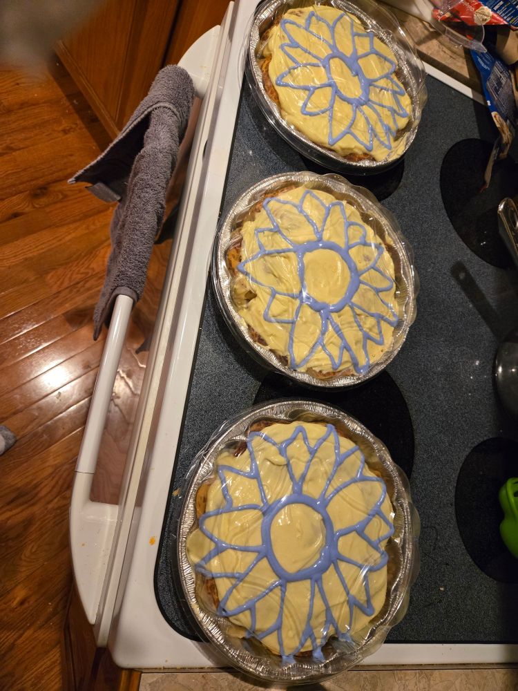 a picture of three round baking pans of cinnamon rolls on a stovetop. the icing is primarily yellow with a blue outline of a sunflower on each.