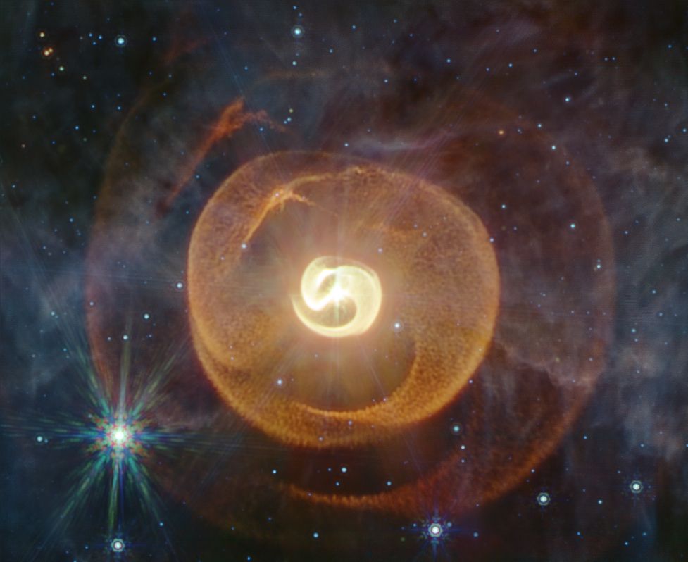 A nebula with a bright star at its center. The nebula has three distinct layers, and the pattern in the layers repeats in a way that implies there are more layers outside the image boundaries, though they would be faint. The shape of the nebula is something like nesting yin yangs, with the center smallest one bright and dense, shining, smooth and golden. The middle one is orange and textured like a sherpa blanket. The largest, outer shell is fainter, and red like an ember. Its texture has softened along with its dissipation into space. There are some small stars speckled around the image, and one annoyingly bright, spiky one that's got too many colors because of the way the light spreads from point sources through the telescope's optics. A pale haze of grayish cloud also covers the scene, but it is sheer and translucent.