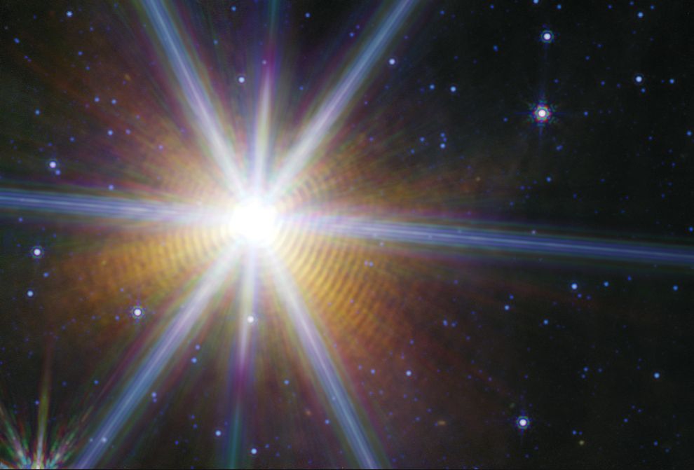 A starry space scene. There are small blue stars scattered randomly but fairly evenly around the image. One bright star dominates the image left of center by several orders of magnitude. It is surrounded by reddish orange shells which visually appear as rings or a very tightly wound spiral. Each shell is almost exactly the same shape as the shell preceding it, resulting in an unusual geometric oddity. The slightly imperfectly circular shapes further lend to the baffling presentation of the image. There are 8 prominent blueish multi-colored diffraction spikes interjecting from the star. They are hard to ignore, but have little meaning beyond indicating that the star was very bright on the detector of the telescope.