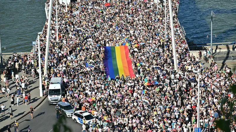Auf dem Foto sieht man die Budapest Pride am heutigen Samstag. Tausende Menschen strömen über eine Brücke und tragen dabei eine riesige Regenbogen-Fahne. 