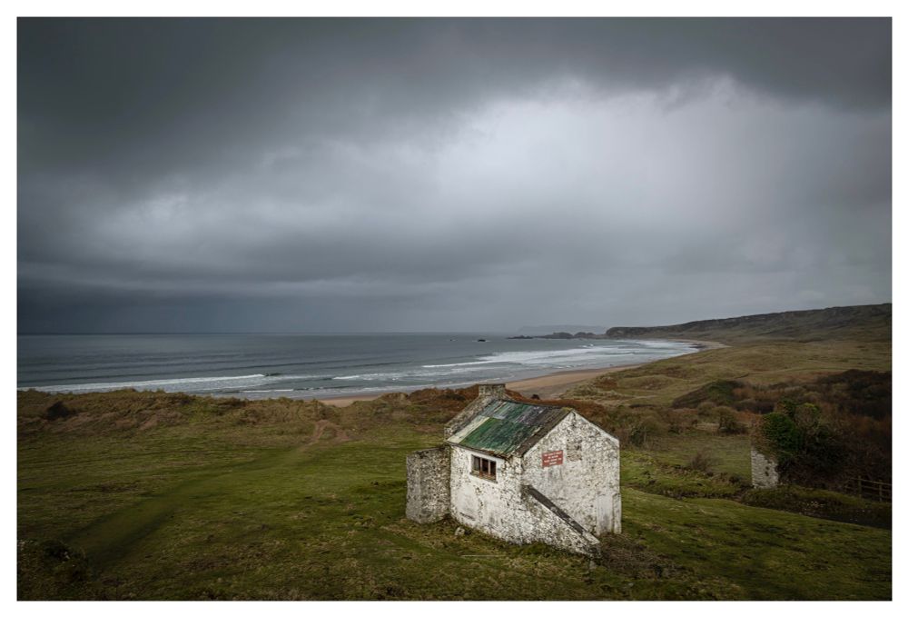 A large curved sandy beach under grey angry overcast skies. A small whitewashed building with a green corrugated iron roof sits in the foreground.