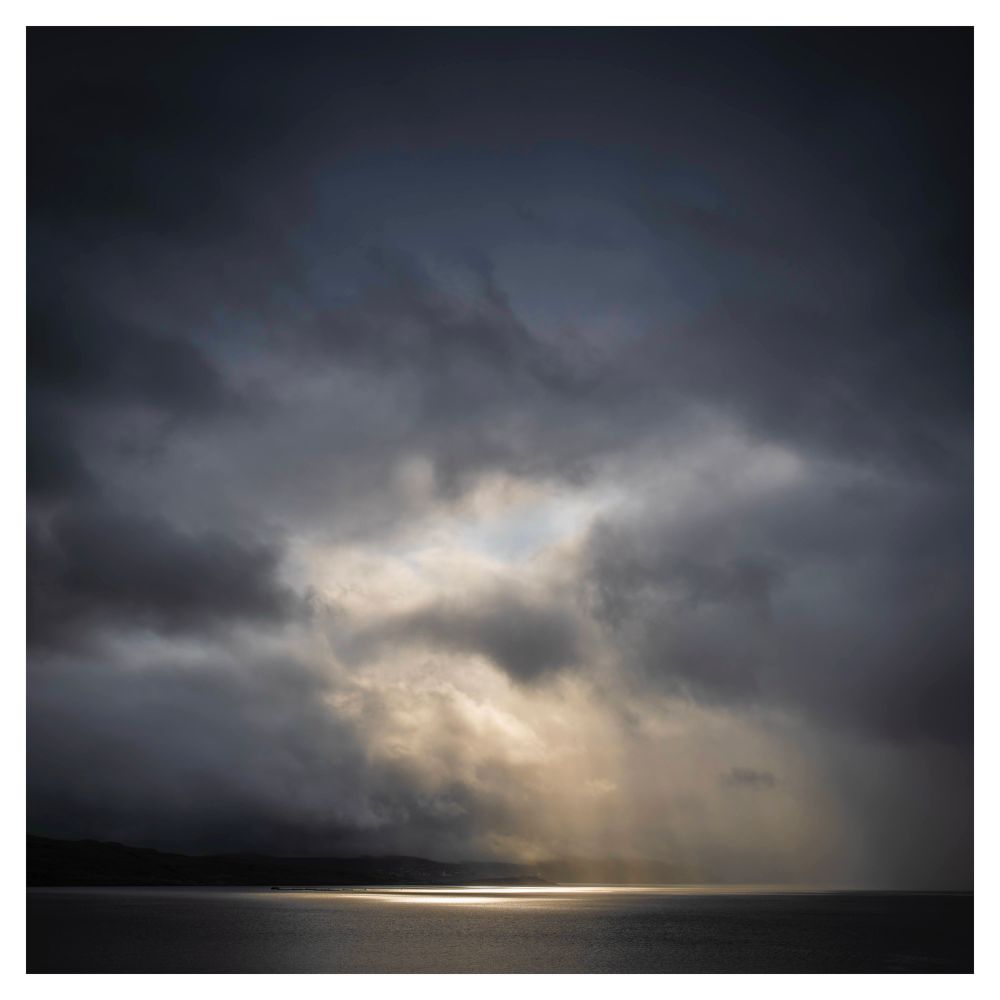 A seascape with heavy foreboding skies, however a break in the clouds lets some light through creating a pool of colour on the sea. From the Faroes.