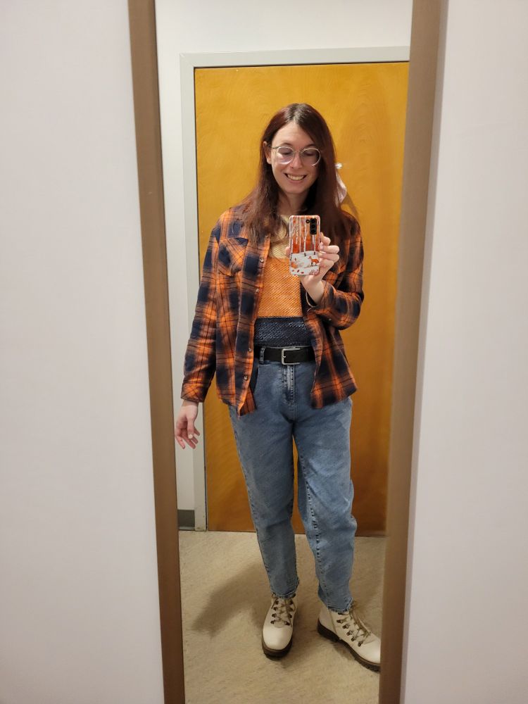 Selfie of Chloe and her whole outfit. Her outfit consists of high waisted baggy jeans that are dark blue, a sweater that is blue, beige, and orange, white boots, and an orange and dark navy blue flannel. She is looking down at the camera to make sure the photo comes out more even than the previous one. 