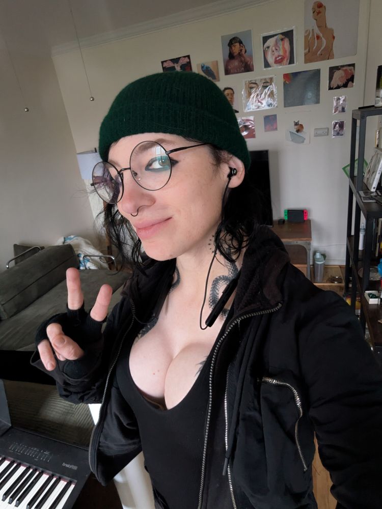 Selfie of a woman in a mostly black techwear look, giving a peace sign. Her cleavage is cleaving cleavefully