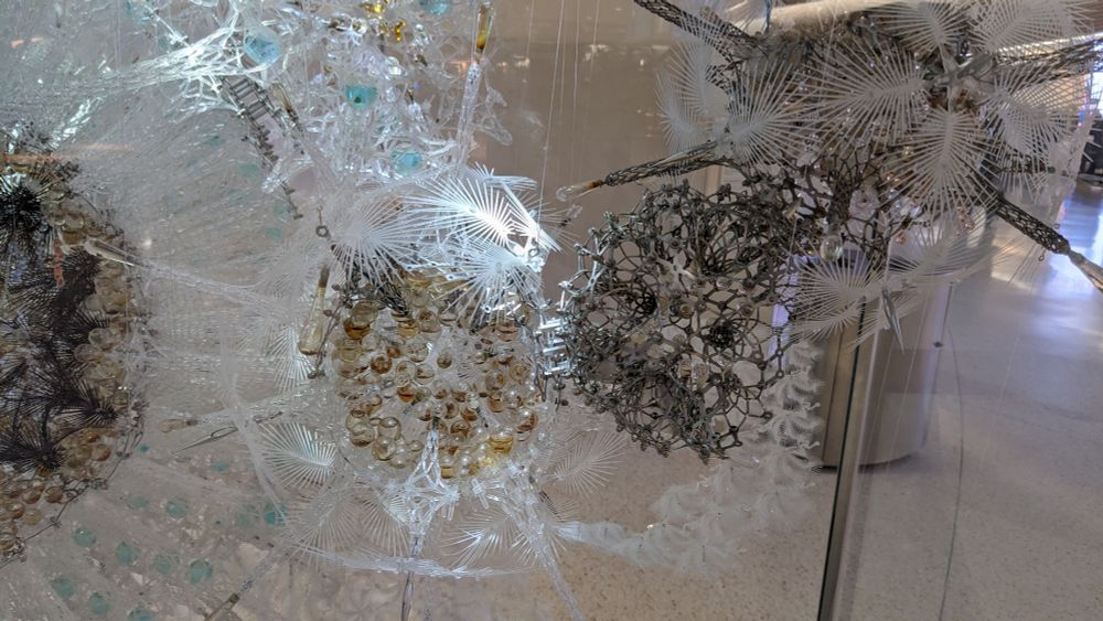 Close up. The art is made of various beads, wires, and feathery plastic pieces assembled into cell-like forms. 