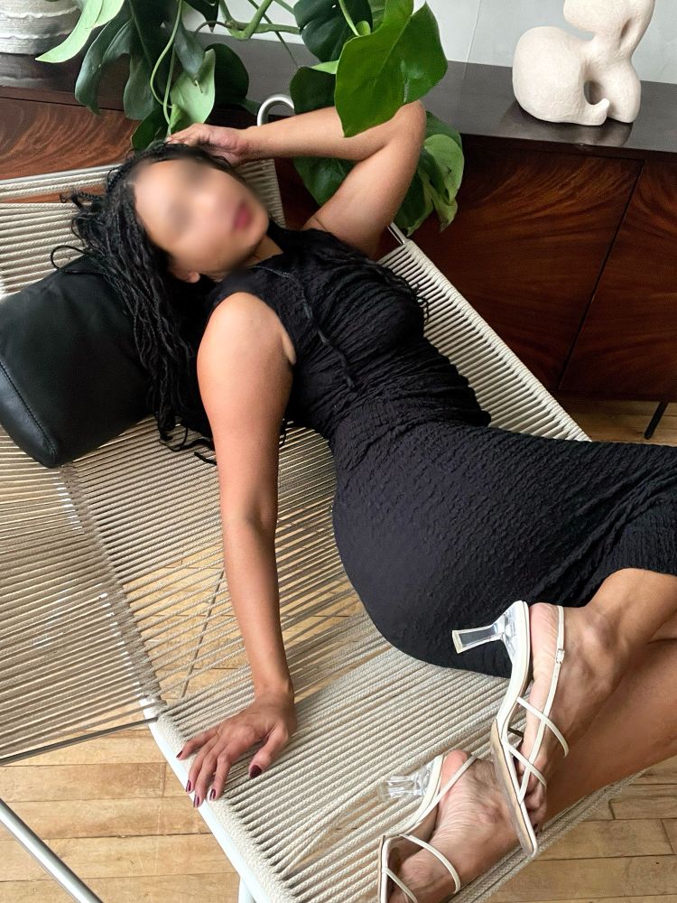 Maya stretches on her side in a creme designer chair. Her body is in an open position. She’s wearing a black textured high neck sleeveless dress with creme colored sandals. There are plants & art behind her on a midcentury wooden dresser. 
