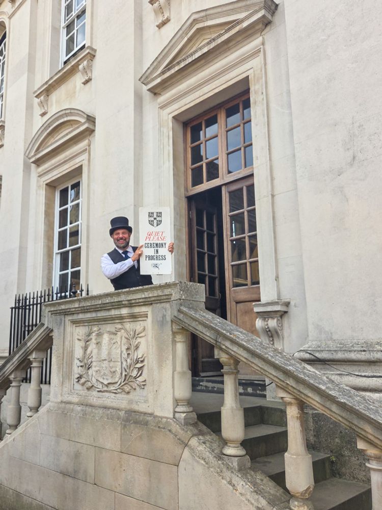 An official standing at the exit of the Senate House in Cambridge holding a sign that says "Quiet please ceremony in progress"