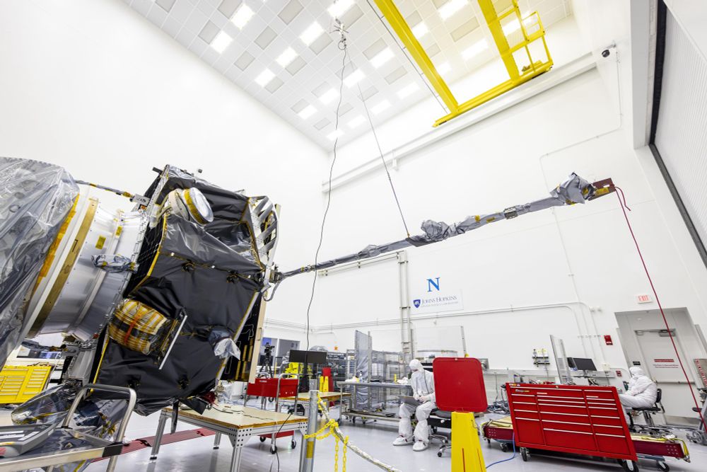 A photograph of the IMAP spacecraft and the MAG magnetometer in the clean room. The spacecraft is wrapped in various insulating/protective fabrics on the left of frame in a brightly lit, white-walled space. The MAG instrument extends from the side of the spacecraft across much of the centre and left hand side of the frame. An engineer sits working in a protective white suit at the bottom of the frame. Credit: NASA/Johns Hopkins/Princeton/Ed Whitman