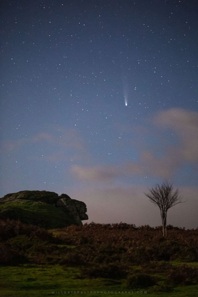 A photograph of a comet over a rocky tor outcrop and moorland landscape lit by moonlight. The comet appears as a fuzzy spot of light two thirds of the way up the frame with a diffuse tail extending above it into a starry moonlit sky. 