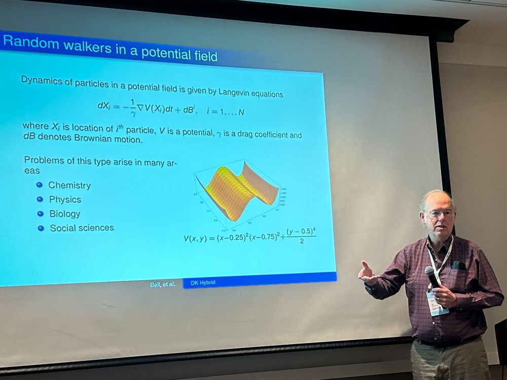 Presenter stands in front of a projection screen displaying a slide titled "Random walkers in a potential field," which includes mathematical equations, a 3D plot, and a list of application areas such as chemistry, physics, biology, and social sciences.